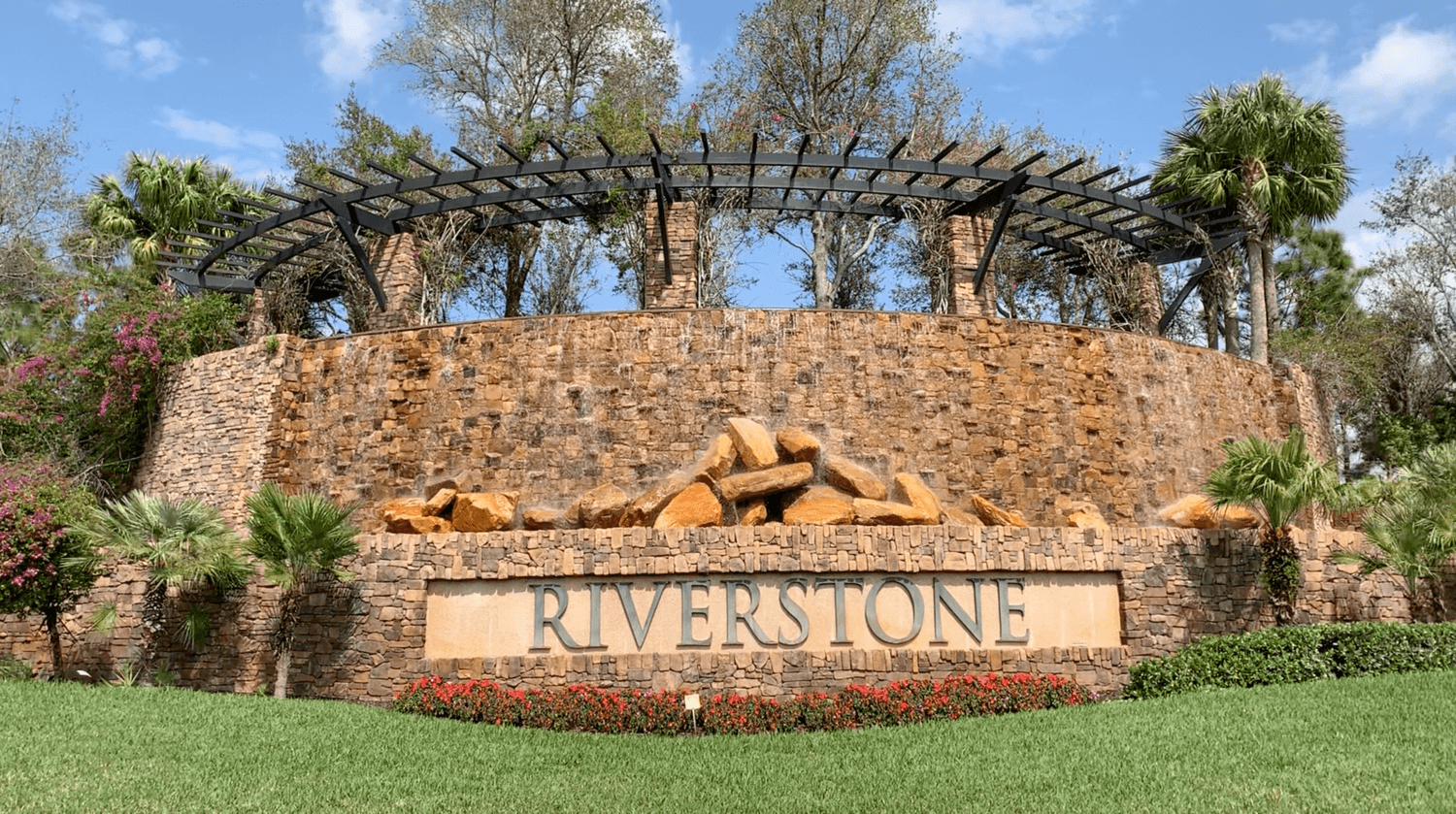 Front entrance sign built of stone and incorporated with tall trees to the Riverstone Naples community.